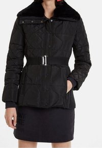 Svart quiltad jacka med bred midja, zippered fickor och krage av konstpäls, bärs över en svart kjol.