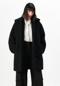 O femeie cu păr drept și închis la culoare poartă o haină oversized texturată de culoare neagră peste o cămașă albă cu nasturi și pantaloni negri cu croială largă, cu mâinile în buzunare.