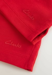 Tela roja con una superficie texturizada, con costuras sutiles y un logo bordado "Clarks" en rojo a juego. No se ven otros detalles.