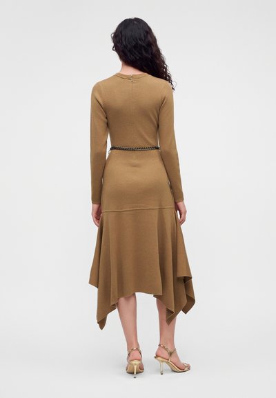 Un mannequin vu de dos porte une robe beige à manches longues et à l’ourlet asymétrique, une fine ceinture chaîne et des escarpins dorés.