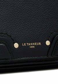 Portafoglio in pelle nera texturizzata con dettagli in metallo dorato. Presenta il logo del marchio impresso "LE TANNEUR 1898" e borchie tonde per un tocco di stile.