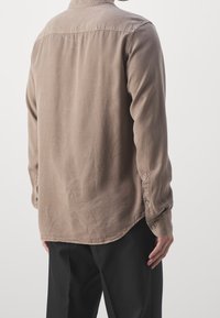 Camisa marrón de manga larga con cuello abotonado y dobladillo curvado. Presenta costuras visibles y botones decorativos en los puños.