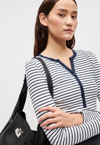 Μπλούζα με μακριά μανίκια σε ριγέ λευκό και ναυτικό μπλε χρώμα με λαιμόκοψη henley με κουμπιά. Μαύρη τσάντα με λαμπερή μεταλλική λεπτομέρεια κρέμεται στον ώμο.