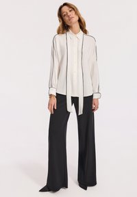 Biała bluzka z czarnym wykończeniem, zapinana z przodu, z wiązanym kołnierzykiem i długimi rękawami, połączona z szerokimi czarnymi spodniami i spiczastymi czarnymi butami.