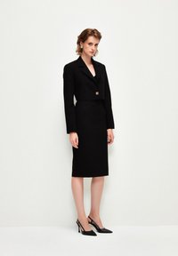 Černý vypasovaný blazer s krátkým designem, zapínání na jeden knoflík a dlouhými rukávy, spárovaný s přiléhavou černou tužkovou sukní a černými lodičkami.