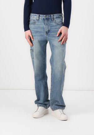 Light blue denim jeans with a relaxed fit, featuring front and side pockets, and subtle fading. Styled with white sneakers.