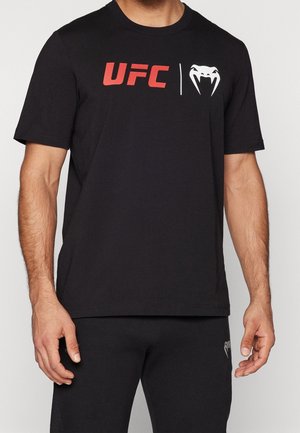 Man draagt een zwart UFC T-shirt met een rood logo en een wit slangenverenteken, gecombineerd met een zwarte broek met een klein slangensymbool.