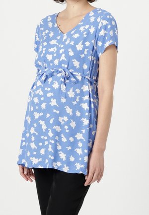 Femme enceinte portant un haut de maternité bleu à motifs floraux avec manches courtes et ceinture à nouer, associé à un pantalon noir.