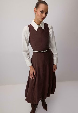 Donna che indossa un vestito marrone senza maniche sopra una camicetta bianca con colletto e polsini smerlati, accessoriato con una sottile cintura a quadri e stivali marroni.