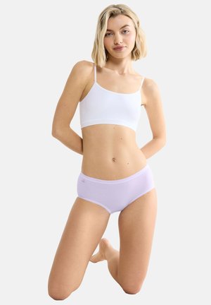 Junge Frau mit blondem Haar kniet, trägt ein weißes, kurzes Tanktop und hellviolette Unterwäsche vor einem schlichten Hintergrund.