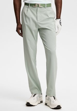 Mężczyzna ubrany w jasnozielone spodnie o prostym kroju, pas w tym samym kolorze, białą koszulę, białą rękawiczkę do golfa oraz białe trampki na białym tle.