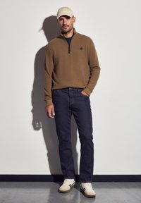 Sudadera con cremallera marrón, vaqueros de mezclilla oscuros, gorra beige y zapatillas blancas con acentos marrones. Diseño sencillo con un acabado de tela texturizada.