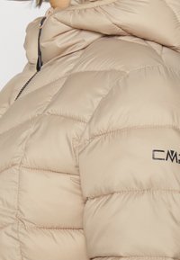 Beige puffjakke med quiltet design, skinnende overflade og et logo på ærmet. Indeholder en hætte og en kontrasterende lynlås.