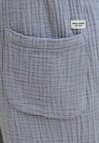 Tissu gris texturé avec une poche avant cousue et une étiquette blanche "JACK & JONES EST. 1990" au-dessus de la poche.