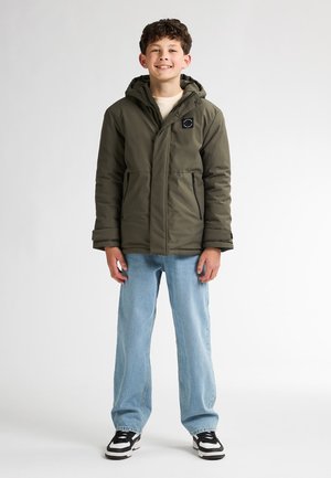 Doudoune vert olive avec capuche, poches zippées, patch logo noir, associée à un jean bleu clair et des baskets noir et blanc.