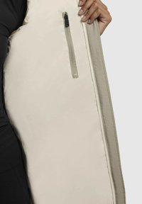 Abrigo aislante beige con una superficie texturizada. Presenta un bolsillo lateral con cremallera y un borde en un gris claro que contrasta a lo largo de los bordes.