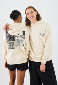 Felpa crema con stampa grafica sulla schiena, polsini e orlo a costine, abbinata a pantaloni corti neri e jeans.