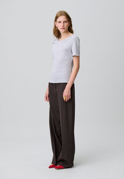 T-shirt grigio chiaro, aderente, con maniche corte, abbinato a pantaloni larghi marroni e scarpe rosse con punta. Anello dorato in evidenza sulla mano.