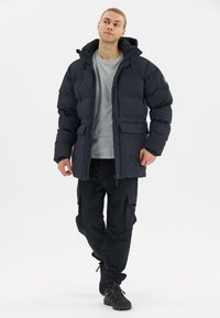 Gepolsterte schwarze Jacke mit Kapuze, mit einem Reißverschluss und mehreren Taschen. Getragen über einem grauen Hemd und kombiniert mit schwarzen Cargo-Hosen.