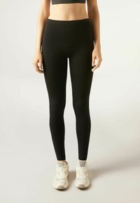 Schwarze Leggings aus dehnbarem Material mit hohem Bund und glatter Textur, kombiniert mit weißen Sportschuhen.