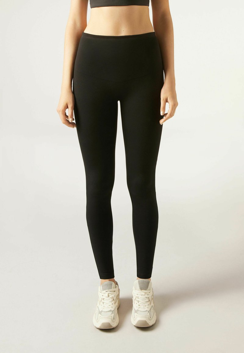 Schwarze Leggings aus dehnbarem Material mit hohem Bund und glatter Textur, kombiniert mit weißen Sportschuhen.