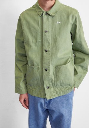 Homme portant une veste verte boutonnée avec le logo Nike sur la poitrine et un jean bleu clair, debout les mains détendues le long du corps.