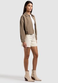 Femme debout portant une veste courte marron, un haut blanc, un short crème et des bottines beiges sur un fond gris clair uni.