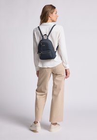 Navyfarbener Stoffrucksack mit zwei Schulterriemen, einer Fronttasche und Reißverschlüssen in goldener Farbe. Getragen über einem weißen Hemd und hellbeigen Hosen.