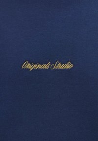 Material bleumarin cu o textură subtilă, având brodat în litere cursive aurii textul "Originals Studio". Design simplu și curat.