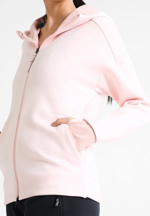 Vrouw draagt een lichtroze hoodie met ritssluiting en geribde manchetten, met handen in de voorzakken, gecombineerd met een zwarte broek.