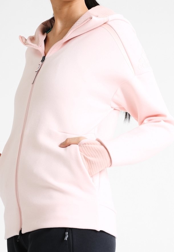 Femme portant un sweat à capuche rose clair zippé avec des poignets côtelés et les mains dans les poches avant, associé à un pantalon noir.