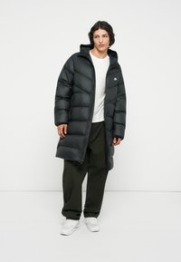 Abrigo de plumas negro con capucha, de longitud larga, con cierre de cremallera y textura acolchada. Lleva puesto sobre una camiseta blanca y se combina con pantalones oscuros.