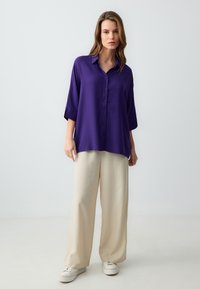 Chemisier violet à manches trois-quarts, associé à un pantalon beige ample et des baskets blanches sur un fond neutre.