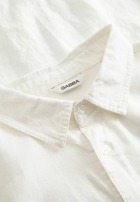 Weißes Baumwollhemd mit klassischem Kragen, ausgestattet mit einem Gabba-Label und zwei Knöpfen. Der Stoff hat ein leicht strukturiertes Erscheinungsbild.