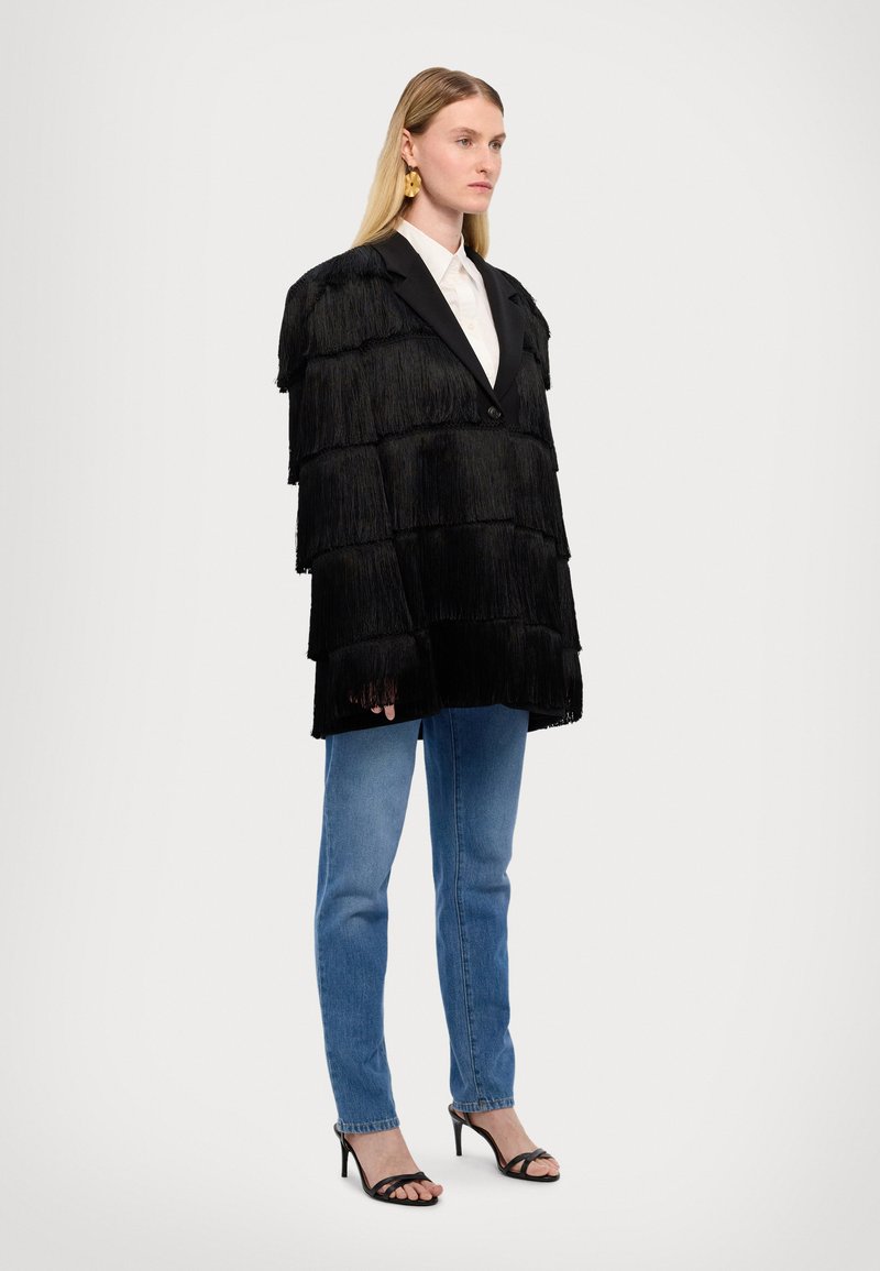 Zwarte fringejacket met een gelaagd ontwerp en reverskraag, gecombineerd met lichtblauwe jeans en zwarte hoge hakken met bandjes. Soepele textuur.