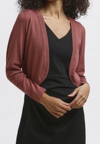 Kvinna i en vinröd cardigan över en svart klänning, som försiktigt justerar cardigans med sina händer. Hon har lockigt hår och en avslappnad hållning.