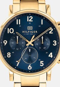 Montre-bracelet dorée avec un cadran bleu, grands chiffres blancs, trois sous-cadrans et un bracelet en métal poli. Présente un logo au centre.