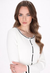White ribbed cardigan with black accents, round neckline, and button closure. Long sleeves feature contrasting black trim at cuffs.