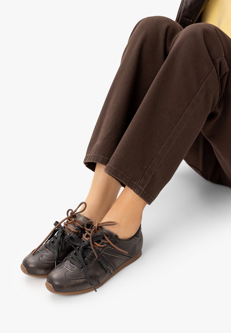 Personne portant un pantalon marron foncé et des chaussures en cuir marron avec des lacets noirs et marron, assise avec les jambes étendues sur un sol blanc.