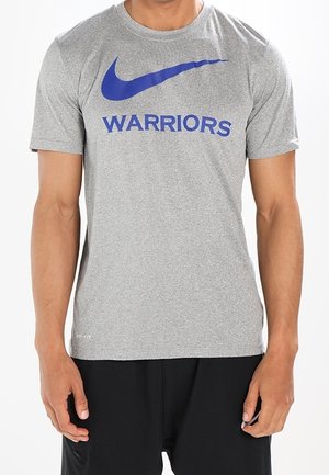 Homme portant un t-shirt gris Nike Dri-FIT avec un swoosh Nike bleu et le texte "WARRIORS", assorti à un short noir.