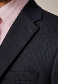 Marineblauwe blazer met textuur en een notch lapel, lichtroze gestippelde shirtkraag zichtbaar eronder, en een discrete pochette als detail.