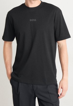 Homme portant un t-shirt noir uni à manches courtes avec le mot « BOSS » brodé en petites lettres au centre de la poitrine.