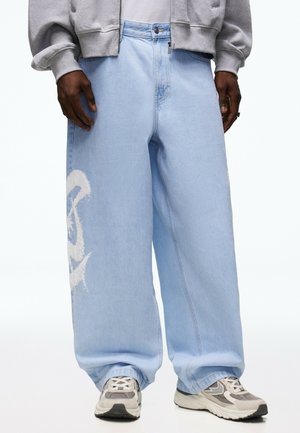 Personne portant un jean large bleu clair avec un motif blanc imprimé sur le côté, un sweat à capuche gris zippé et des baskets grises.