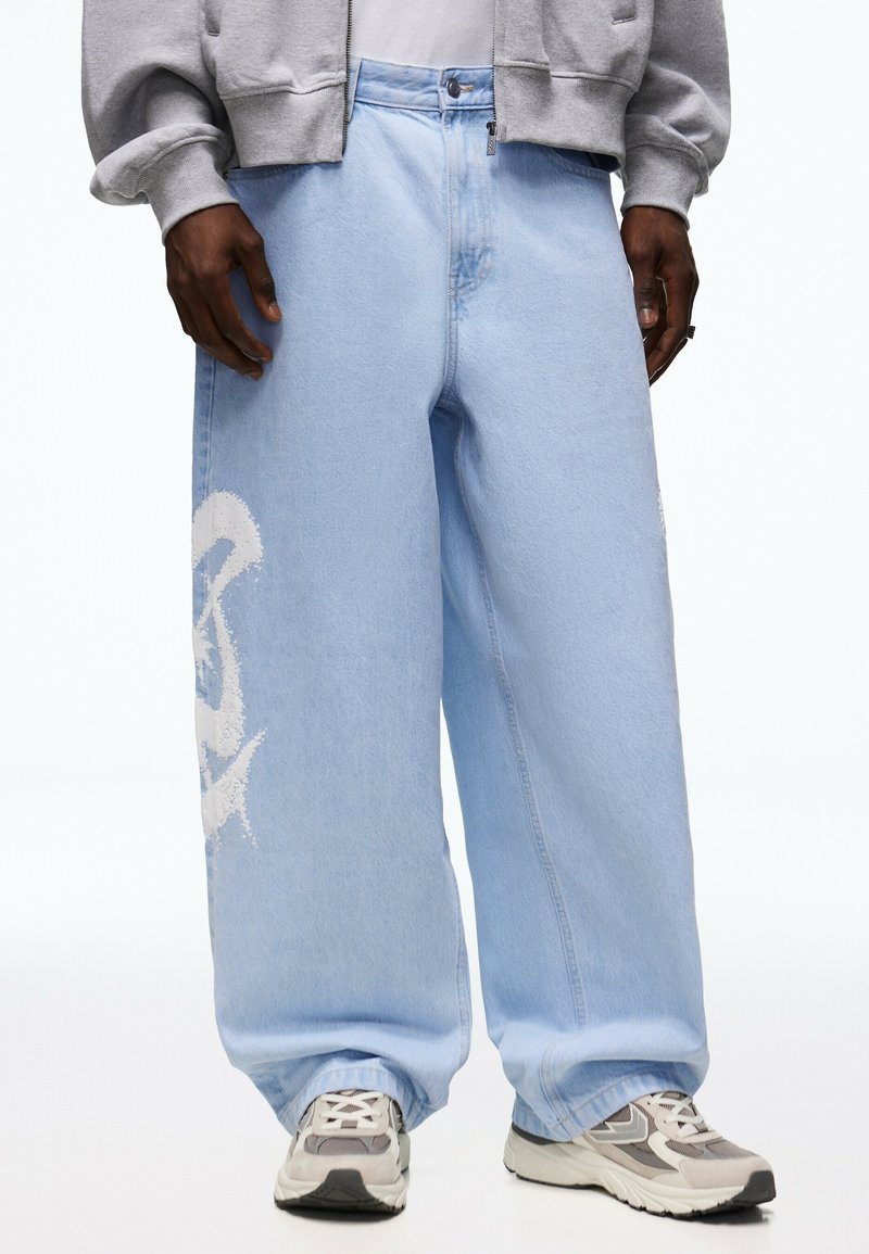 Personne portant un jean large bleu clair avec un motif blanc imprimé sur le côté, un sweat à capuche gris zippé et des baskets grises.