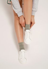 Białe sneakersy z gładkiej skóry, z płaskimi sznurowadłami i zaokrąglonym noskiem. Połączone z zielonymi, prążkowanymi skarpetkami do kostek. Widoczny odcień skóry na nogach.