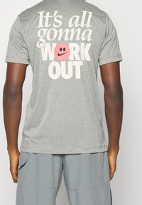 Šedé tričko s krátkým rukávem, které obsahuje velký potisk s textem "Všechno to dobře dopadne," doplněné o růžovou smajlíkovou tvář, vyrobené z měkké tkaniny.