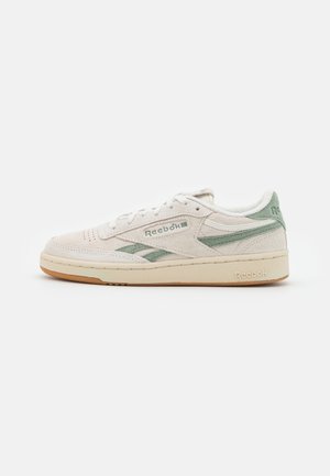 Low-top Reebok sneaker in beige suède met groene accenten, geperforeerde neus, witte veters en gum rubberen zool op een witte achtergrond.
