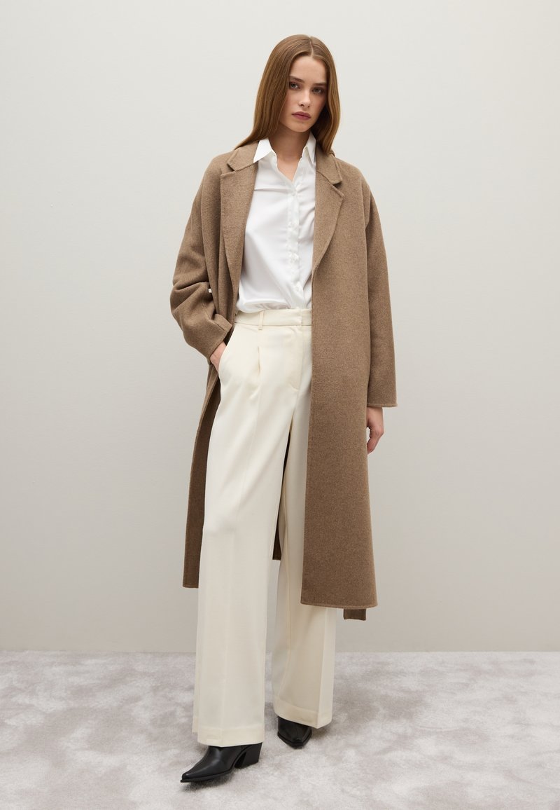 Manteau en laine brun avec des revers larges, porté sur une chemise blanche et un pantalon large crème. Bottines noires à talons pointus. Textures lisses.