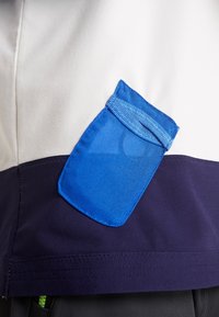 Biała i granatowa kurtka z niebieską siateczkową kieszenią. Kieszeń ma elastyczny górny brzeg i gładką teksturę materiału.