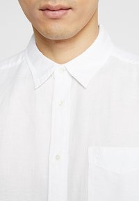 Camicia bianca in lino con colletto button-down, caratterizzata da una texture sottile e una piccola tasca sul petto con logo ricamato.